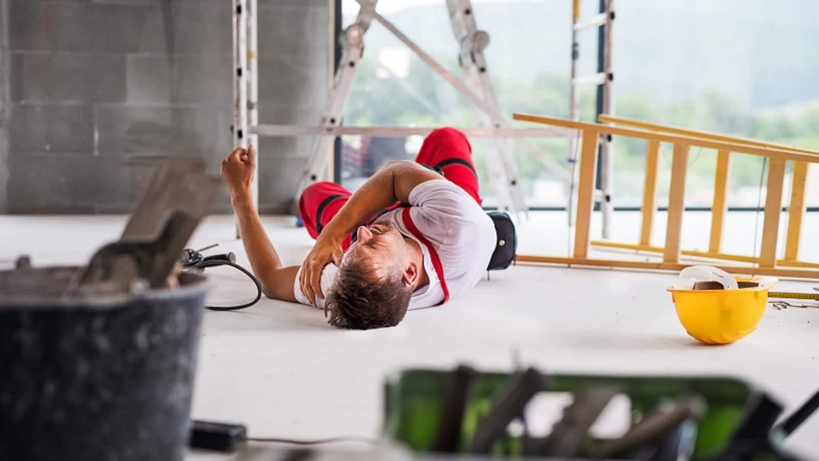 An injured worker clutches his shoulder while he lies on the ground beside a fallen ladder.