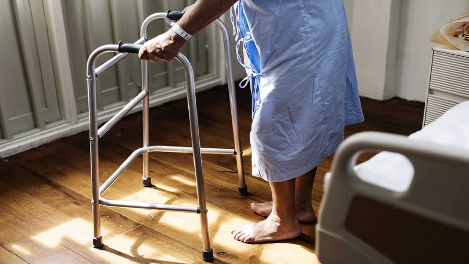 An elderly person in a hospital gown stands near a window holding onto their walker.