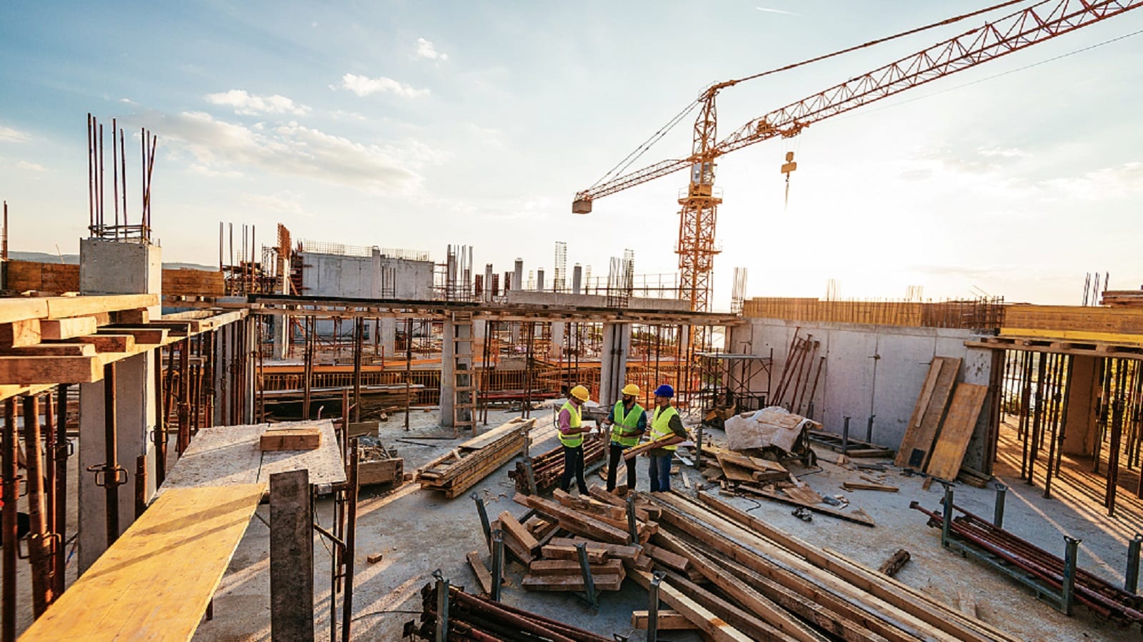 Three construction workers wearing safety vests and helmets are standing on a building site surrounded by scaffolding, wooden beams, and construction equipment, with a crane in the background.