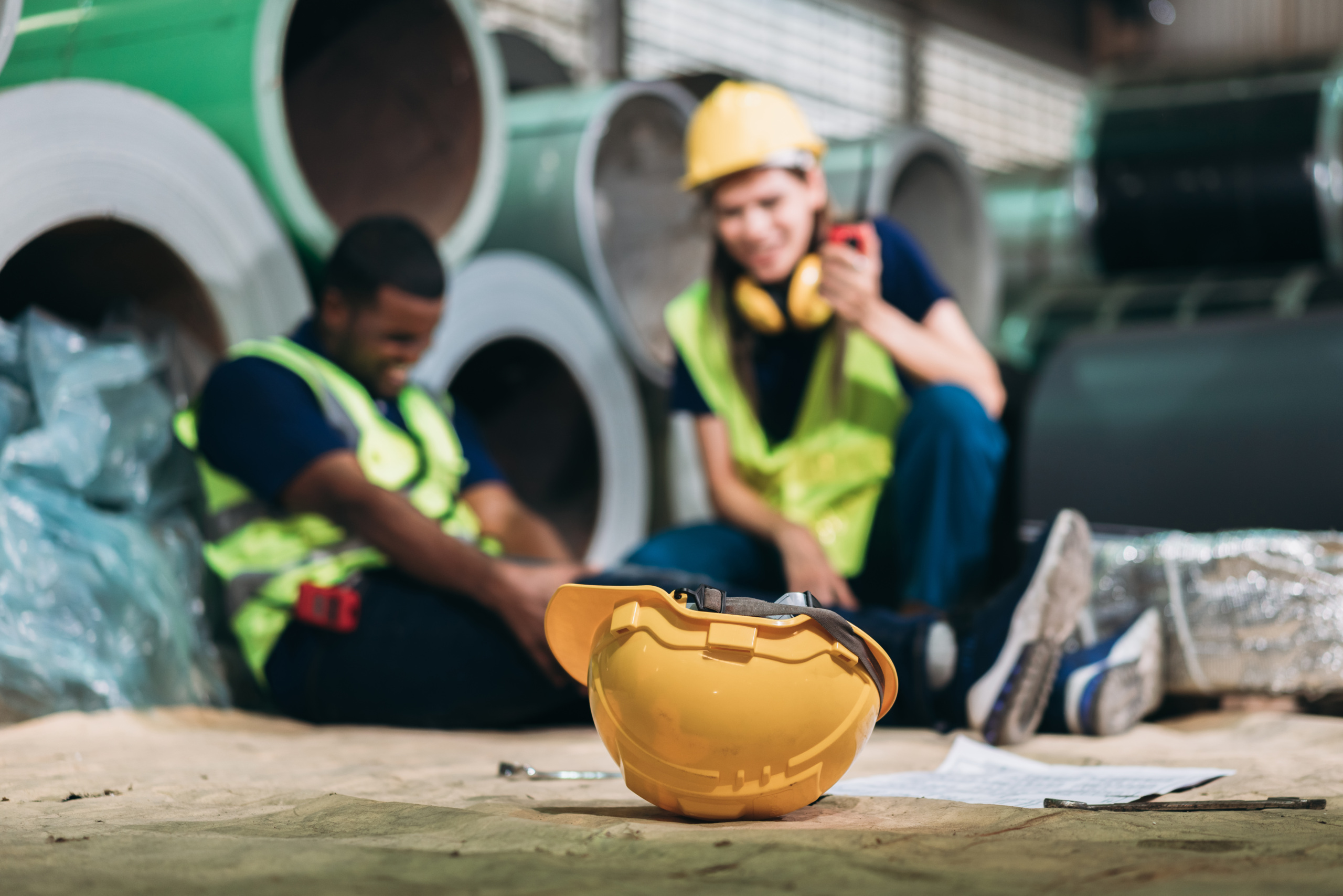 An injured construction worker holding his leg while his coworker radios for help, with a yellow construction hat in the foreground.